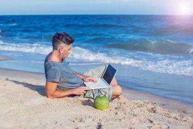 Genç adam sahilde dizüstü bilgisayarla oturuyor.