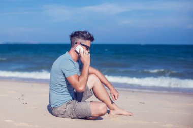 Genç adam sahilde akıllı telefon kullanıyor.