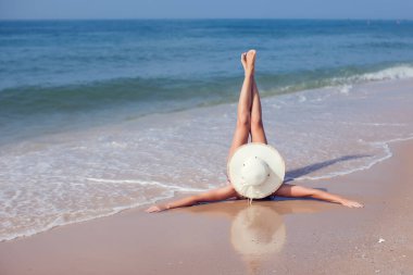 Hasır şapkalı genç bir kadın tropikal bir plajda uzanmış ince bacakları uzatıyordu. Arka planda mavi deniz. Yaz tatili kavramı.