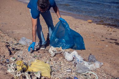 Kumsaldaki çöpleri toplamak için büyük çantalı mavi tişörtlü gönüllü bir adam. Çevre kirliliği kavramı