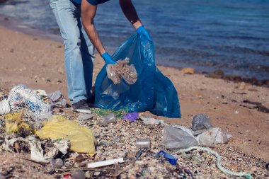 Kumsaldaki çöpleri toplamak için büyük çantalı mavi tişörtlü gönüllü bir adam. Çevre kirliliği kavramı