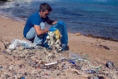 Kumsaldaki çöpleri toplamak için büyük çantalı mavi tişörtlü gönüllü bir adam. Çevre kirliliği kavramı