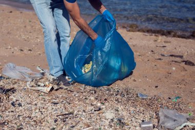 Kumsaldaki çöpleri toplamak için büyük çantalı mavi tişörtlü gönüllü bir adam. Çevre kirliliği kavramı