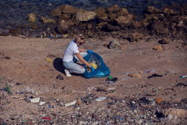 Beyaz tişörtlü, büyük mavi çantalı, plajda çöp toplayan gönüllü bir kadın. Çevre kirliliği kavramı