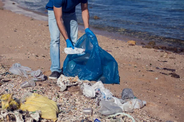 Kumsaldaki çöpleri toplamak için büyük çantalı mavi tişörtlü gönüllü bir adam. Çevre kirliliği kavramı