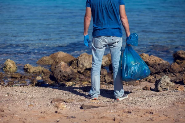 Kumsaldaki çöpleri toplamak için büyük çantalı mavi tişörtlü gönüllü bir adam. Çevre kirliliği kavramı