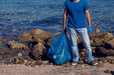 Kumsaldaki çöpleri toplamak için büyük çantalı mavi tişörtlü gönüllü bir adam. Çevre kirliliği kavramı