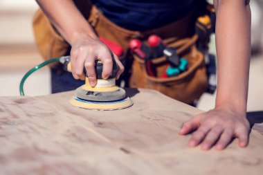 Kısa sarı saçlı, güler yüzlü genç bir kadın ahşap yüzeyi zımparalıyor.. 