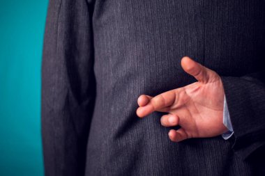 Businessman in suit keeping crossed fingers sign behind his back in front of black background