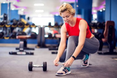 Woman in red top with short blond hair tying shoelaces before training in gym. People, fitness and lifstyle concept