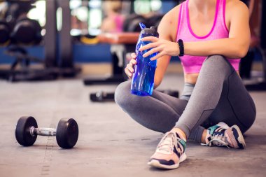 Woman in a pink top and short blond hair holding blue bottle with water resting in the gym. People, fitness and health care concept