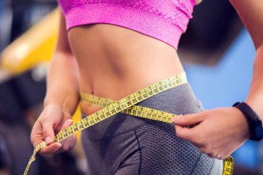 Woman in pink top and gray leggins measuring her waist in the gym. People, fitness and health care concept