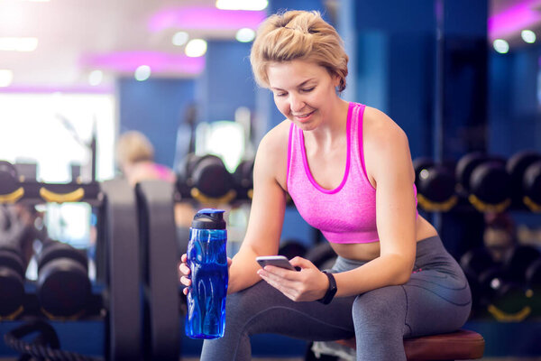 Woman in pink top with short blond hair resting in the gym, sitting on bench with bottle of water and smartphone. People, fitness and health care concept