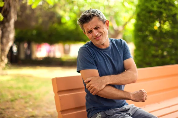 肘の痛みを屋外で感じる男 男が公園のベンチに座っている 医療の概念 — ストック写真