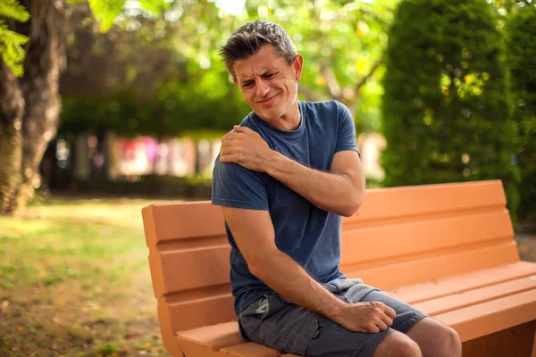 Man with shoulder pain sitting on the bench in the park. Healthcare and medicine concept