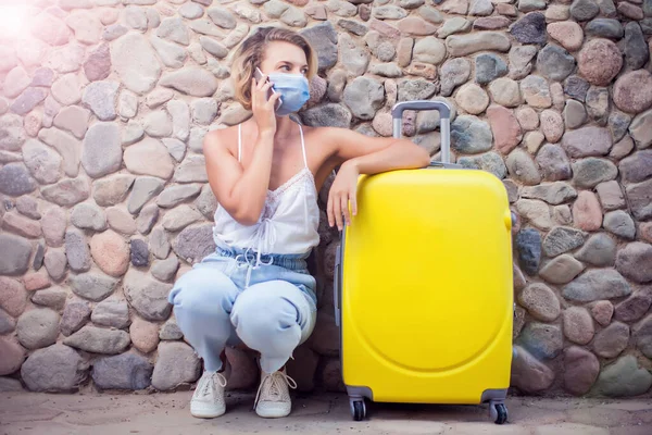 Woman with luggage wearing medical face mask and using smartphone outdoor. Travel and coronavirus concept