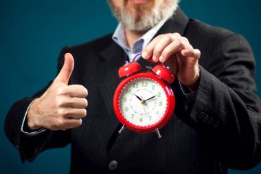 Sakallı iş adamı kırmızı alarm saati tutuyor ve baş parmağını kaldırıyor. Zaman yönetimi kavramı