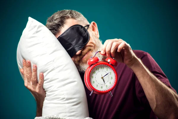 Surprised man in red t-shirt and sleep mask on head holding red alarm clock and white pillow. Lifestyle and bed time concept