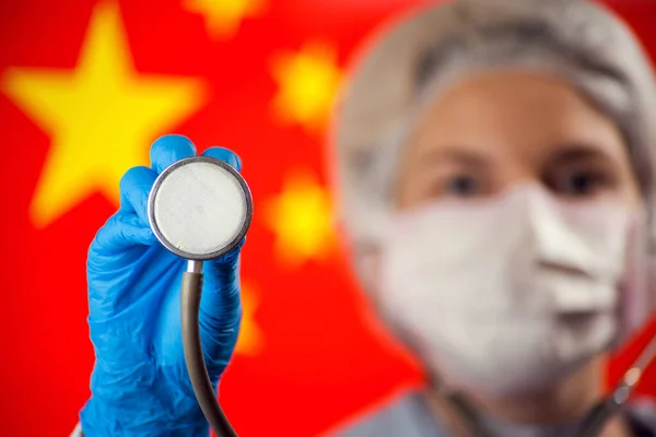 Female doctor in medical mask and blue gloves holding phonendoscope in front of flagf of China. Coronavirus, epidemic and healthcare concept