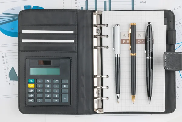 Deník, kalkulačka a tři pera leží na pozadí diagramu — Stock fotografie