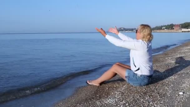 La mujer se sienta en la playa del mar de Azov y arroja piedras al agua . — Vídeos de Stock