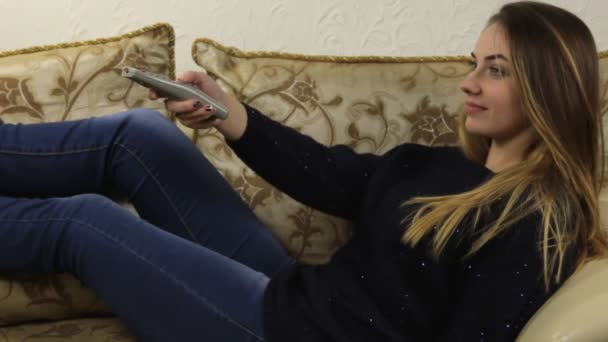 Ung vacker flicka liggande på soffan, dricka ur en mugg och hanterar fjärrkontroll — Stockvideo