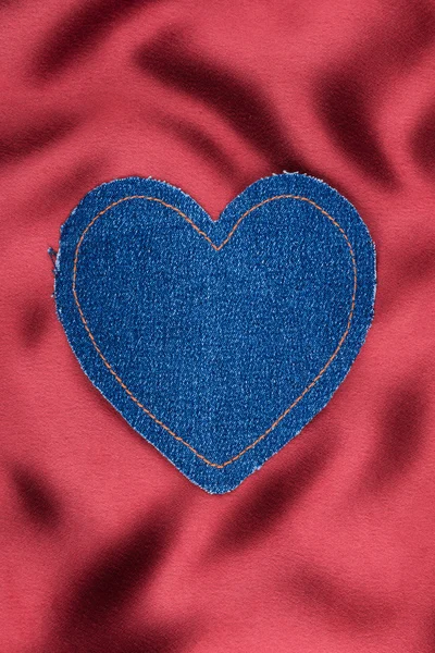 Heart made of denim fabric with yellow stitching on red silk — Stock Photo, Image