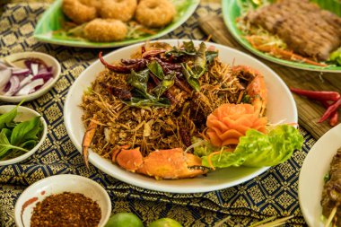 Yengeç pençeli bir Tayland yemeği, doğranmış sebzeler, kurutulmuş biberler ve yeşil otlar. Oyulmuş sebze çiçeğiyle süslenmiş ve fesleğen, baharat, limon ve desenli kumaş üzerine diğer Tayland yemekleriyle çevrili..