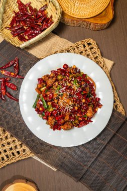 Kurutulmuş kırmızı ve yeşil biberlerle çevrili, susam tohumlarıyla süslenmiş baharatlı kızarmış tavuk tabağı, sade dokuma hasırında servis edilir..