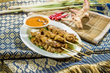 Kızarmış satay şişleri yeşil yapraklar üzerinde servis edilir. Fıstık sosu, limon otu, kırmızı biber ve geleneksel dokuma kumaşında galangal ile çevrili..