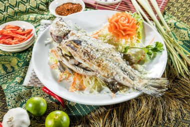 Doğranmış lahana ve havuçla servis edilen ızgara bir balık, oyulmuş havuç çiçeği ve otlarla süslenmiş, Tayland baharatlarıyla çevrili..