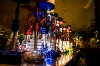 Yansıtıcı bir yüzeye yerleştirilmiş parlak bazlı ve metal saplı renkli nargileler. Bitkiler ve sicim ışıkları gece atmosferini güçlendirir.