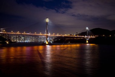 Hong Kong 'daki Tsing Ma Köprüsü' nün gece manzarası, arka planda canlı bir şehir silueti ile aydınlanmış ve suya yansıtılmış..