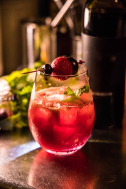 Yuvarlak bir bardakta servis edilen, sıcak ışıklandırmayla vurgulanmış, böğürtlen şişi ve nane filizli kırmızı bir kokteyl..
