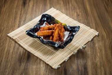 Kırmızı sosla kaplanmış baharatlı tavuk kanatları, kağıt kaplama ve bambu paspaslı siyah seramik kasede servis ediliyor..