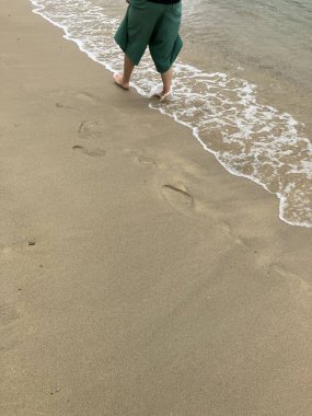 Yeşil şortlu bir insan kıyı boyunca yalın ayak yürüyor, kumlarda ayak izleri bırakıyor. Dalgalar yavaşça içeri giriyor..