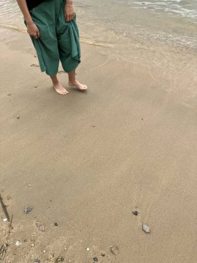 Yeşil pantolonlu ve siyah tişörtlü bir adam sahilin kenarında çıplak ayakla duruyor, sakin ve derin düşüncelere dalmış..