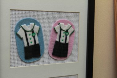 Çerçevelenmiş bir tekstil sanat eserinde, ayrıntılı aksesuarlarla renkli arka planlara monte edilmiş okul üniformalarına benzeyen iki adet keçeli kıyafet yer alıyor..