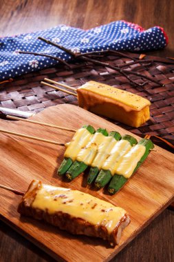 Eritilmiş peynirle kaplanmış üç şişe ızgara bamya, et ve yumurta, ahşap bir tahtada mavi bezle servis edilir..