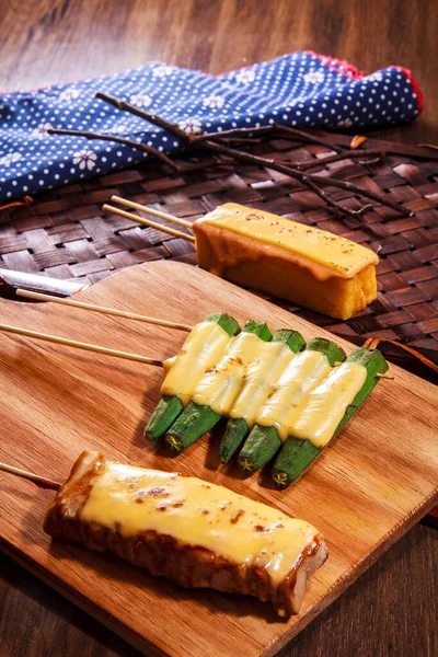 Eritilmiş peynirle kaplanmış üç şişe ızgara bamya, et ve yumurta, ahşap bir tahtada mavi bezle servis edilir..