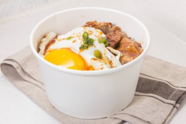 Biftek dilimleri, kızarmış yumurta ve yeşil soğan garnitürlü buğulanmış pirinçle dolu beyaz bir kase..