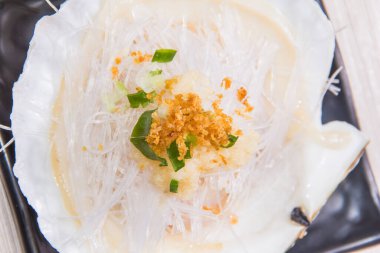 Üzerinde vermicelli, sarımsak kıymalı ve siyah tabakta yeşil soğan garnitürü olan bir midye kabuğu..