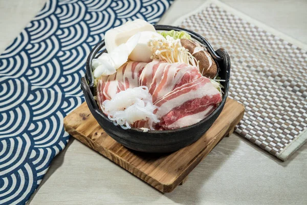İçinde domuz dilimleri, tofu ve mantar içeren Shabu Shabu malzemeleriyle dolu siyah bir tencere..