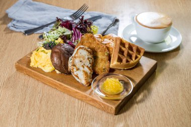 Çırpılmış yumurta, waffle, ızgara tavuk, mücver ve kahveli salata içeren ahşap bir tahta..
