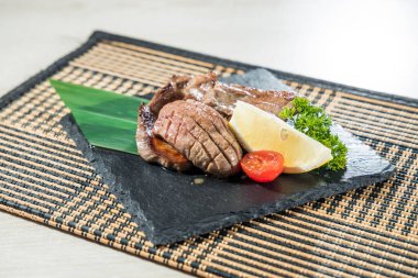 Izgara sığır dili dilimleri limon dilimi ve kiraz domatesli siyah bir levhada servis edilir..