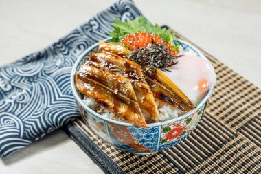 Üstünde ızgara yılan balığı dilimleri, rafadan yumurta ve balık yumurtası olan bir pirinç kasesi desenli bir kasede servis edilir..