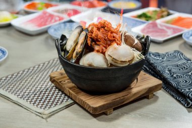 A black pot filled with assorted seafood including clams and mussels, kimchi, mushrooms, and vegetables.