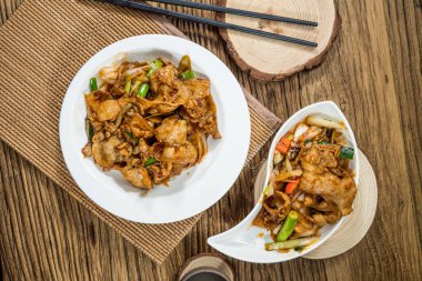 Baharatlı soslu yeşil soğan ve sebzelerle pişirilmiş kızarmış domuz dilimleri, ahşap bir masada beyaz seramik tabaklarda servis edilir..