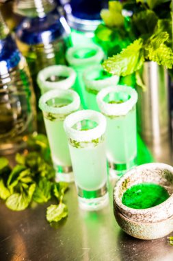 Şeker jantlı shot bardaklarında, taze nane yaprakları ve şişelerle çevrili yeşil naneli kokteyller, canlı bir bar ortamı yaratıyor..