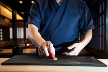 Donanma üniformalı bir suşi şefi bir parça Nigiri 'yi minimalist bir ortamda siyah bir kesim tahtasına yerleştiriyor..